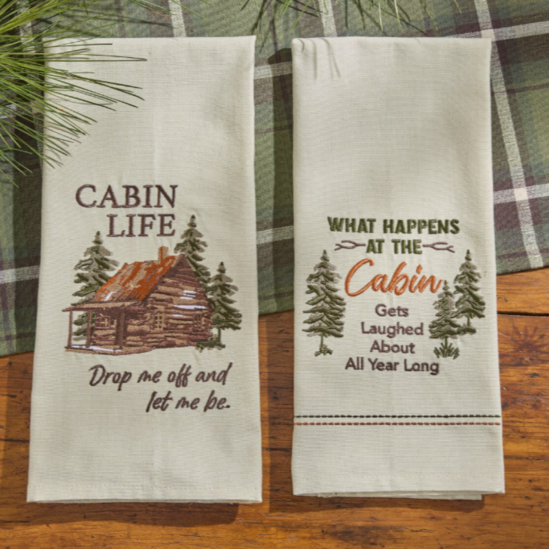 Two embroidered tea towels with cabin-themed text and graphics on a wooden surface.