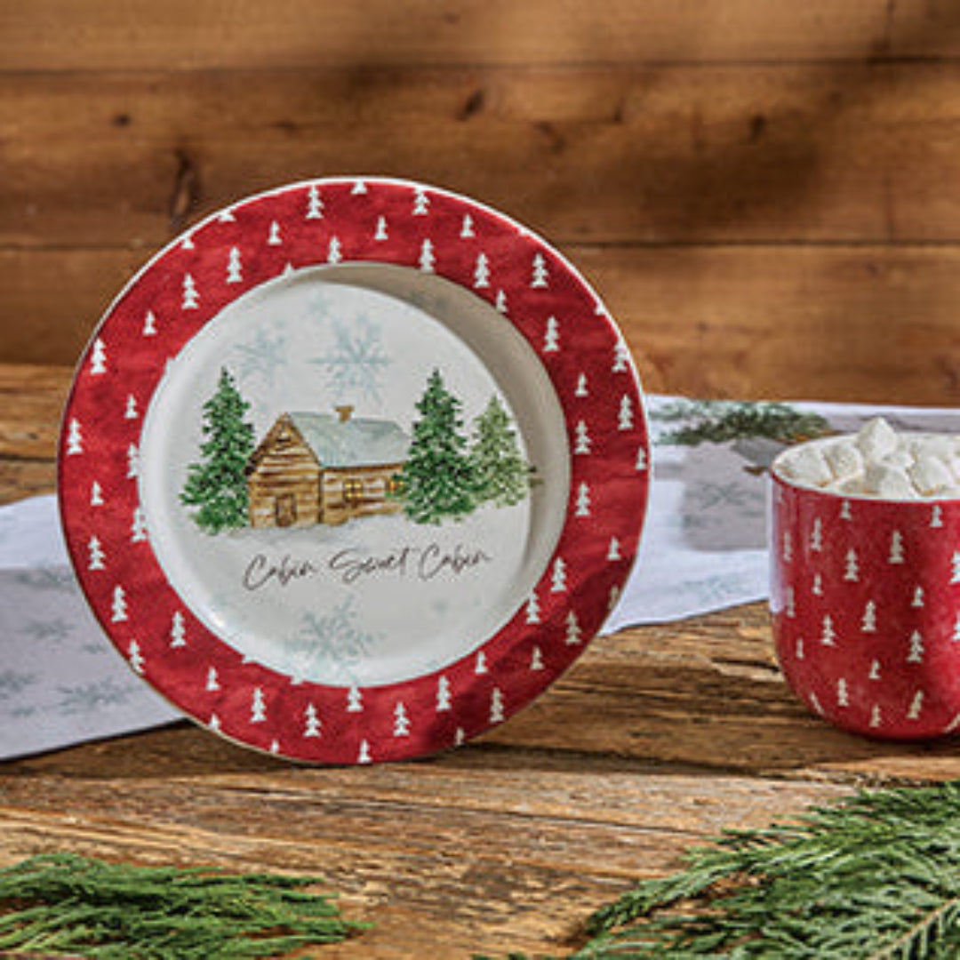 Red and white Christmas-themed mug and plate set on a wooden surface with a cabin and tree design.