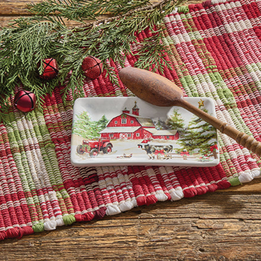 Decorative spoon rest with a farm scene on a red and green woven mat with a wooden spoon.