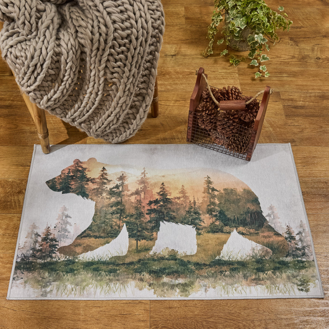 A rectangular area rug with a bear silhouette filled with a forest scene, styled on a wooden floor with a chunky knit blanket, a basket of pinecones, and a potted plant nearby.