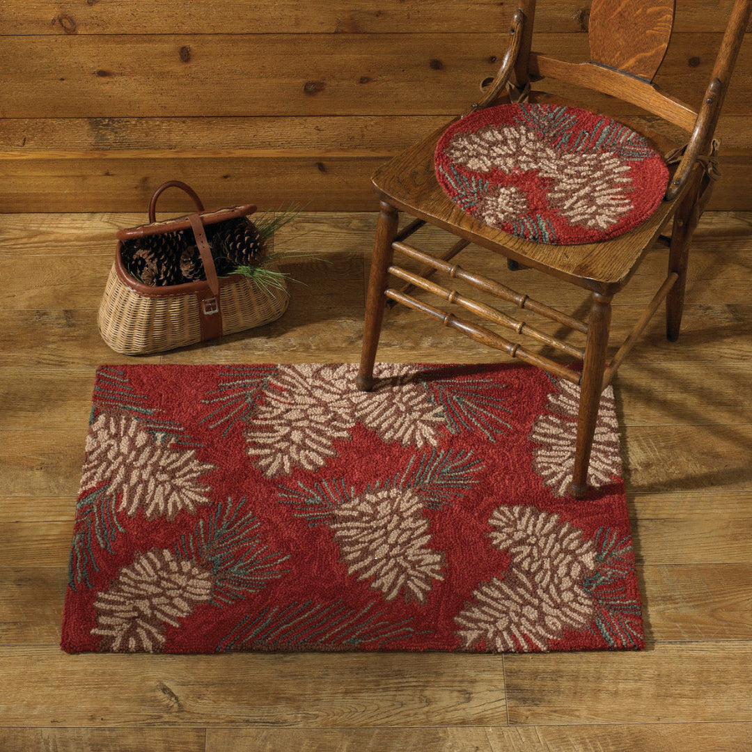 A rectangular red area rug with a large pinecone and pine needle pattern, styled on a rustic wood floor next to a wooden chair with a matching round cushion and a woven basket.