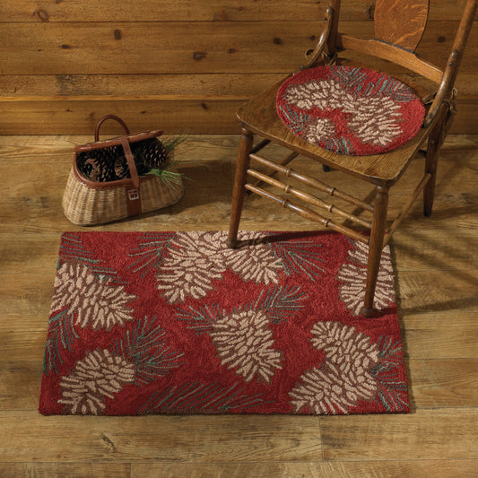 A rectangular red area rug with a large pinecone and pine needle pattern, styled on a rustic wood floor next to a wooden chair with a matching round cushion and a woven basket.