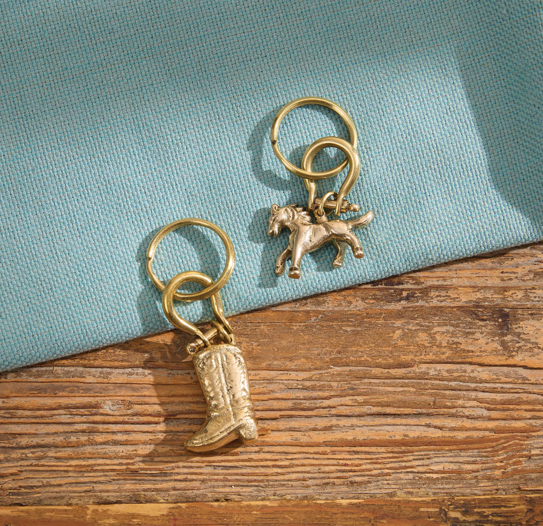 Keychains with split rings are displayed on a rustic wooden surface with a folded light blue fabric backdrop. One charm is a cowboy boot and the other is a horse silhouette.