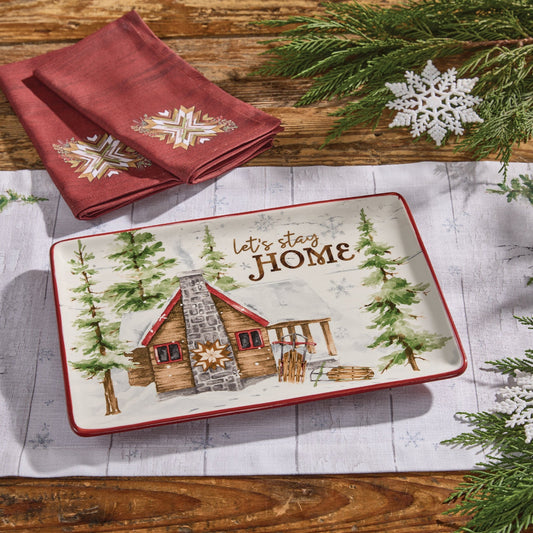 Rectangular platter with a cabin design and 'Let's stay HOME' text, surrounded by Christmas decor on a wooden surface.