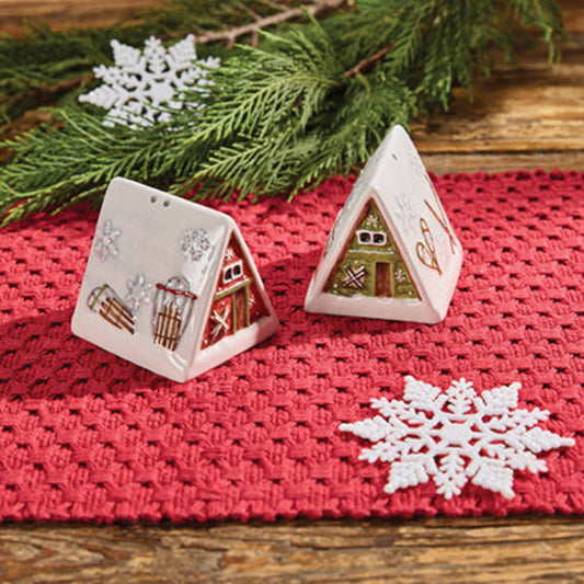 Two decorative gingerbread house salt and pepper shakers on a red woven mat with snowflakes and greenery.