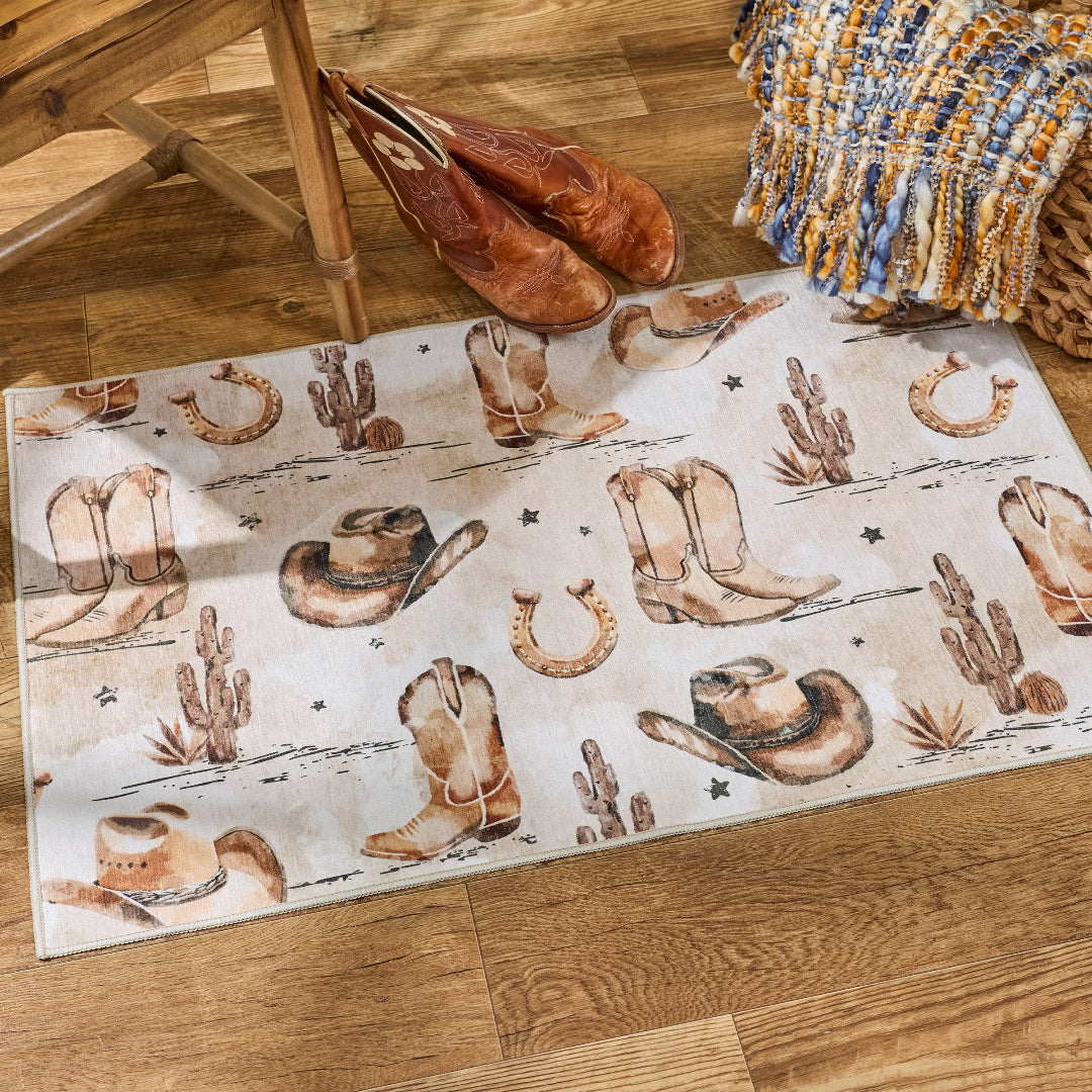 A western-themed area rug with a desert and cowboy motif, styled on a wooden floor next to a wooden chair, brown leather boots, and a blue and yellow throw blanket.