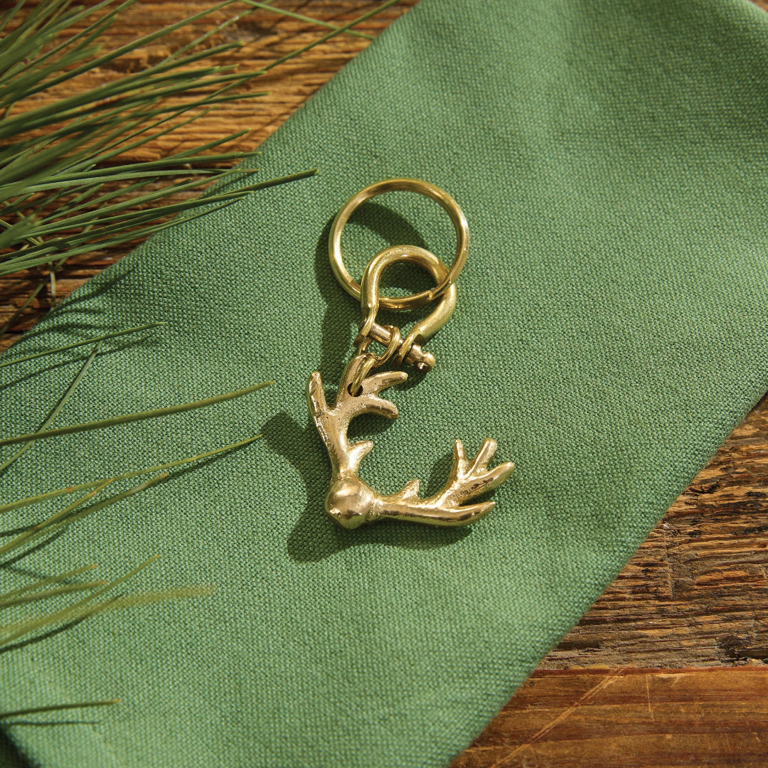 A keychain with a deer head charm resting on a textured green fabric surface, with pine needles and a wooden tabletop visible in the background.