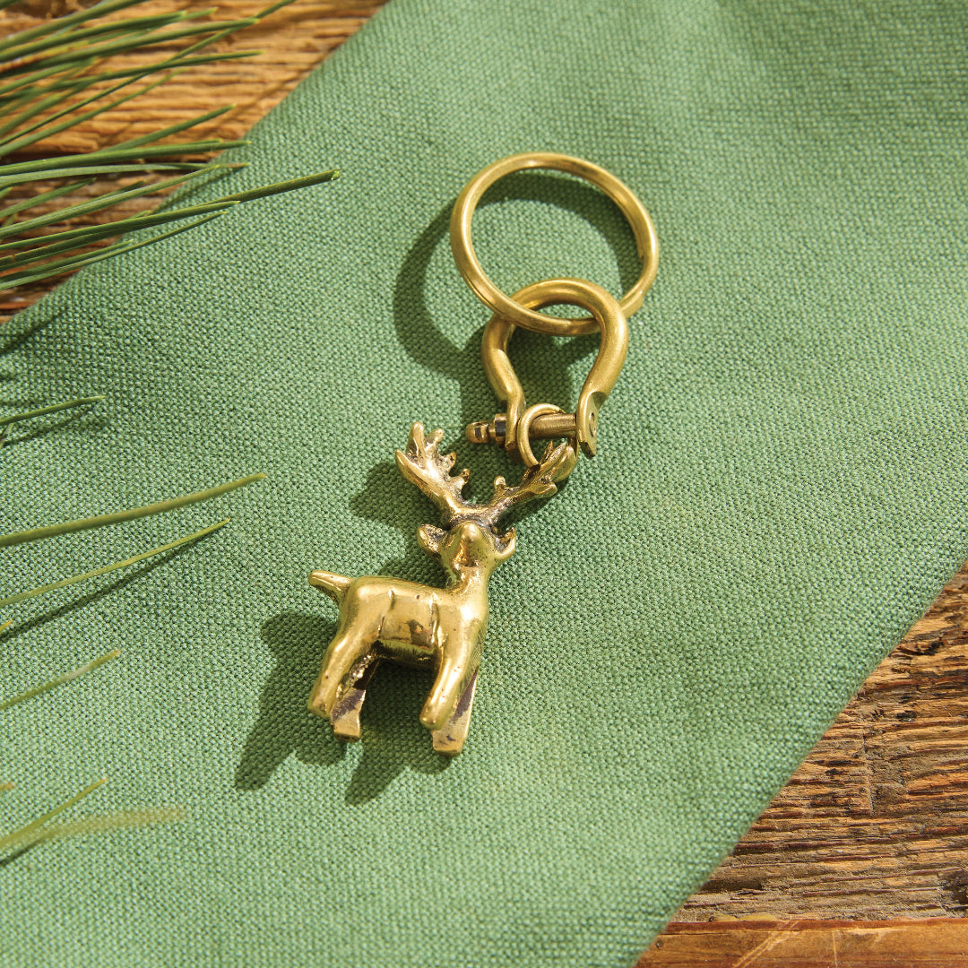 A deer keychain, resting on a textured green fabric surface with pine needles and a wooden background.