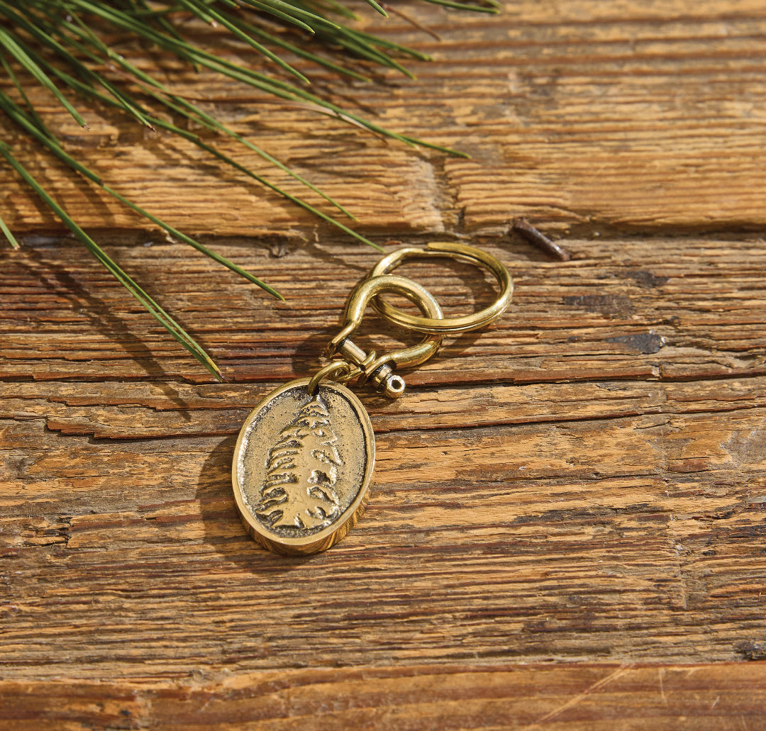 A keychain with an oval pendant featuring a Fir tree design, resting on a rustic wooden surface with pine needles in the corner.