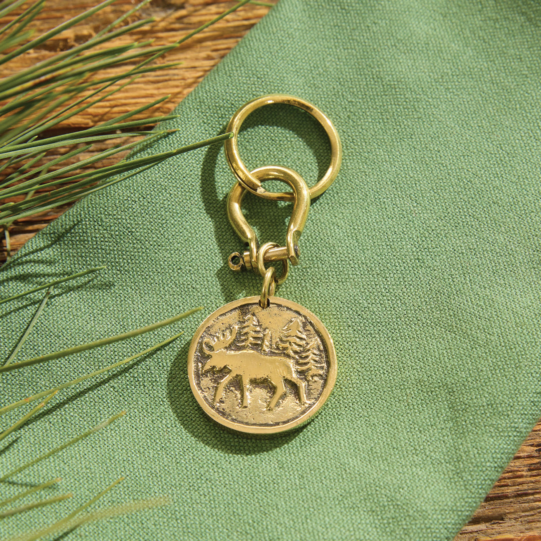 A keychain with a round coin charm featuring a Moose and tree design, resting on a textured green fabric with pine needles and a wooden surface.