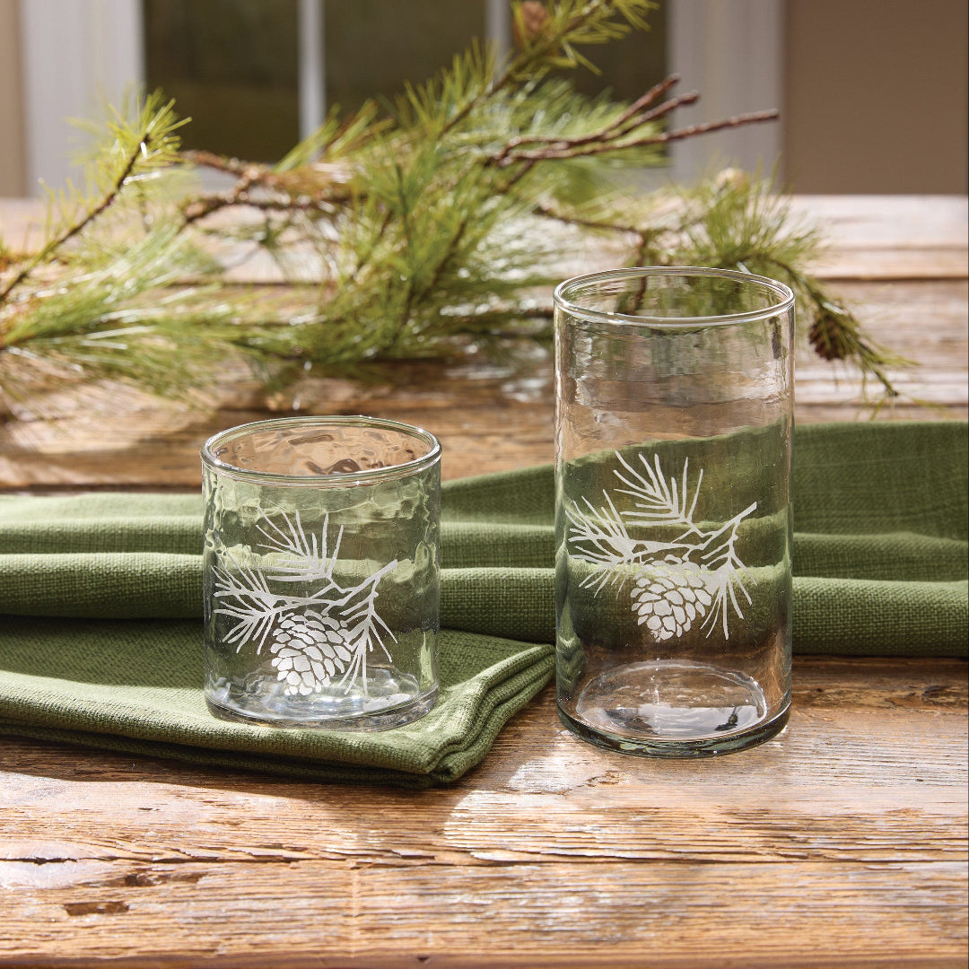 Two clear glass tumblers with white pinecone and pine branch etched designs, displayed on a green cloth napkin on a rustic wooden table with pine branches in the background.