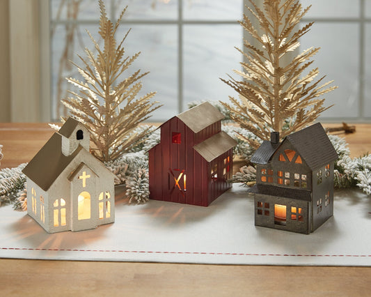 Three small tea lit houses on a table with decorative trees in the background. Christmas Decor
