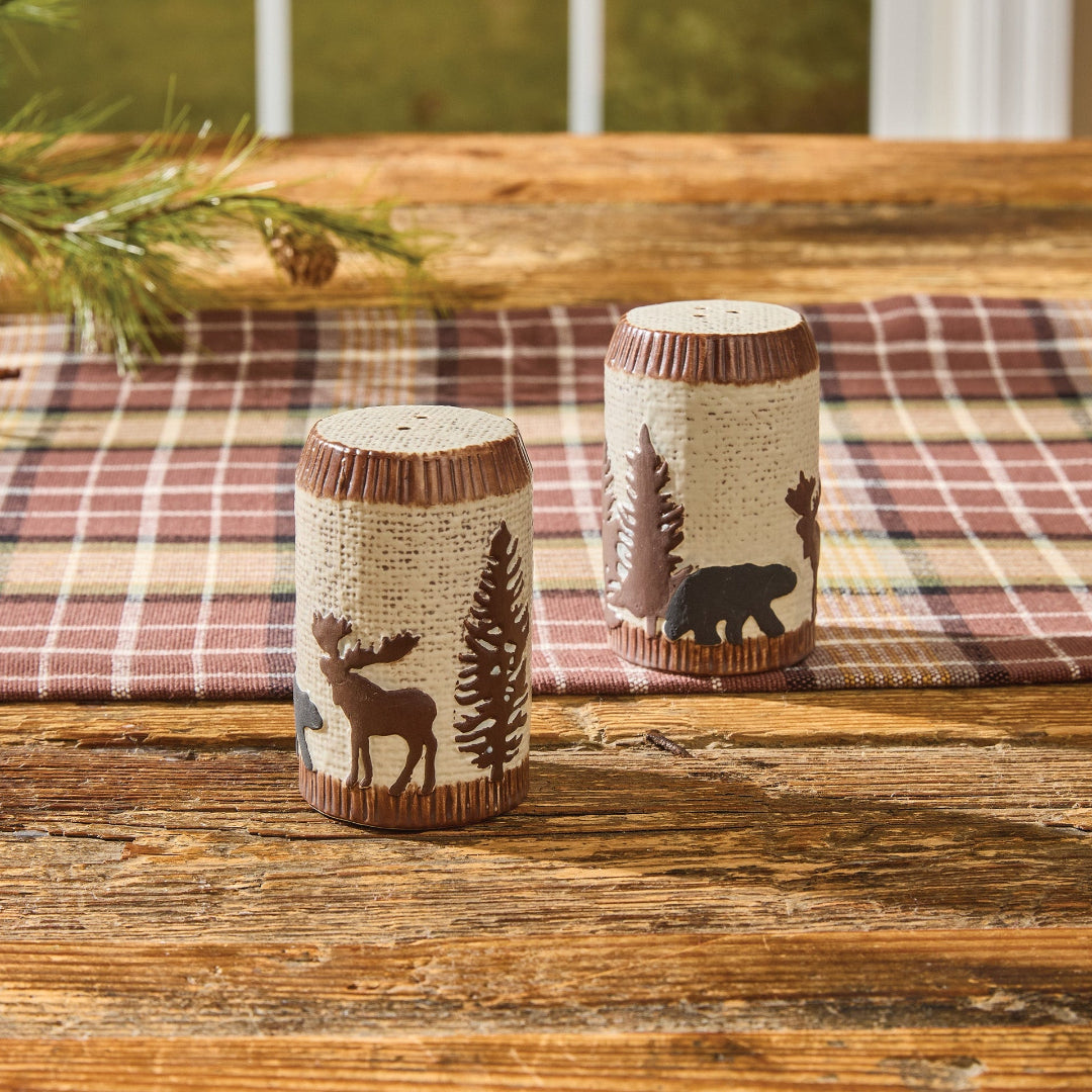 Two decorative Salt & Pepper Shakers with Moose, Bear and nature designs on a wooden surface with a plaid table runner in the background.