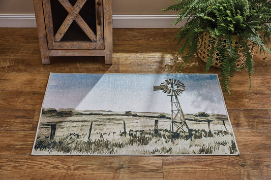 A rectangular area rug featuring a ranch-style landscape print with a windmill, fence, and dry grass, styled on a brown wooden floor next to a rustic wooden cabinet and a potted fern.