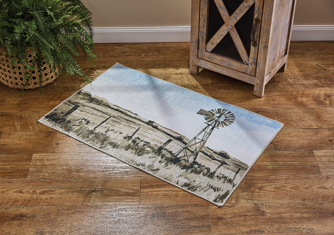 A rectangular area rug with a western farm scene print featuring a windmill and fence, shown on a hardwood floor next to a potted plant and a rustic wooden cabinet.